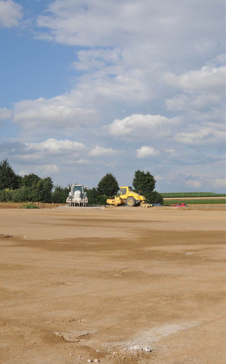 The site is cleared, flattened by bulldozers and ready for development. Building area in the country. Farmland had to yield, the soil will be sealed. The ground has been levelled and prepared for rising a factory or new homes. Wide, empty space. Blue cloudy sky. Trees, bushes, highway and construction machinery in the distance. Horizontal orientation.